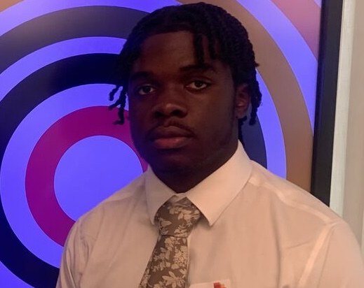 Kingsley, wearing a white shirt and a patterned tie, standing in front of a purple and pink concentric-circle display.